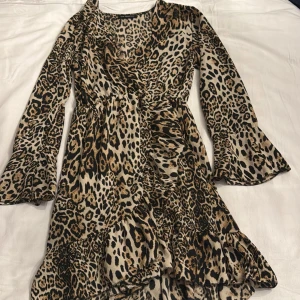 Leopardmönstrad omlottklänning - Trendig leopardmönstrad omlottklänning med volangdetaljer. Klänningen har långa ärmar och en v-ringad hals. Perfekt för att sticka ut med ett djärvt mönster.