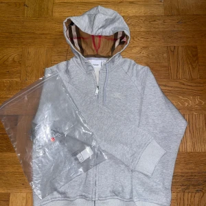 Grå hoodie från Burberry - Säljer en stilren grå hoodie från Burberry med dragkedja och broderad logga på bröstet. Hoodien har en klassisk rutig insida i huvan och ribbade muddar vid ärmslut och nederkant. Perfekt för en avslappnad look. Pris kan diskuteras vid snabb affär!!