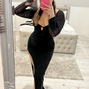 Svart långklänning med glittriga ärmar - Elegant svart långklänning med djup urringning och glittriga långärmade detaljer. Klänningen har en hög slits och en liten öppning framtill. Perfekt för en glamorös kväll.