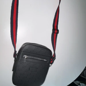 Svart axelväska med mönster - Snygg svart axelväska med präglat mönster och en justerbar axelrem i rött och blått. Väskan har en dragkedja framtill för enkel åtkomst till dina saker. Perfekt för att bära det nödvändigaste med stil.