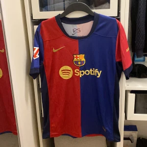 FC Barcelona fotbollströja  - Säljer en officiell FC Barcelona fotbollströja från Nike. Tröjan har spelarnamnet 'Lamine Yamal' och nummer 19 på ryggen. Perfekt för alla Barça-fans. Tröjan är i nytt skick och är i storlek S.