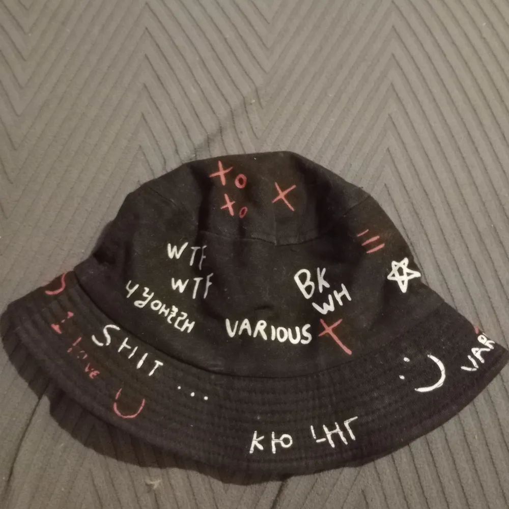 Cool svart bucket hat med olika texter och symboler i vitt och rött. Perfekt för en edgy stil och attityd. Passar bra till avslappnade outfits.. Asusteet.