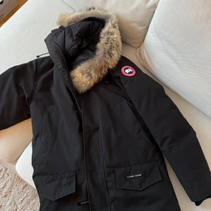 Svart parkajacka med pälsdetalj - Säljer en svart parkajacka HERR JACKA från Canada Goose med pälsdetalj på huvan. Jackan har en klassisk design med två stora fickor framtill och en dragkedja. Perfekt för kalla vinterdagar.  Äkta finns stämpel och serie nummer på jackan ?köpt från Johnells 2019 , helt ny , rökfri och djurfri .