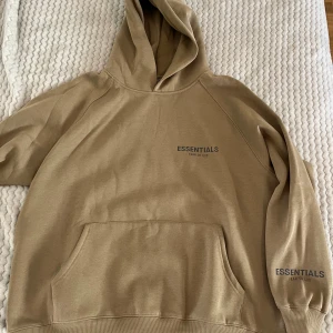 Beige hoodie från Essentials Fear of God - Säljer en stilren beige hoodie från Essentials Fear of God. Den har en klassisk design med en stor ficka framtill och en bekväm huva. Perfekt för en avslappnad look. Har dock aldrig använt den för det passar inte mig.