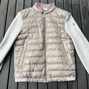 Grå moncler cardigan  - Säljer en snygg grå cardigan från Moncler. Den har en quiltad front med dragkedja och mjuka grå ärmar. Perfekt för höst och vår! Jackan har en stilren design med en liten logga på ärmen. Passar både till vardags och lite finare tillfällen.