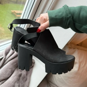 Svarta chunky heels - Snygga svarta chunky heels med hög klack och grov sula. De har en öppen tå och justerbart spänne runt ankeln. Perfekta för att ge lite extra höjd och stil till din outfit. Fint skick, använda endast fåtal gånger men behövs torkas av. Stl 39 