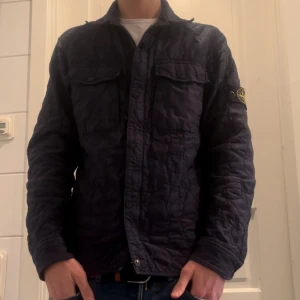 Mörkblå quiltad jacka från Stone Island - Säljer en snygg mörkblå quiltad jacka från Stone Island. Jackan har långa ärmar och knappar framtill. Perfekt för höst och vår med sin stilrena design och bekväma passform.