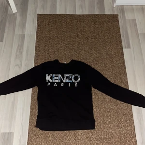 Svart Kenzo Paris tröja - Säljer en svart tröja från Kenzo Paris med deras ikoniska logga i vitt och mönstrat tryck på framsidan. Tröjan har långa ärmar och en klassisk rund halsringning. Perfekt för en stilren look.