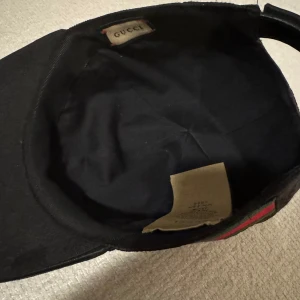 Svart keps från Gucci - Snygg svart keps från Gucci med diskret mönster och klassiska gröna och röda ränder på sidan. Justerbar rem baktill för perfekt passform. Perfekt för att ge din outfit en lyxig touch. Inget kvitto eller box då den är köpt av en kompis. Äkta och i otroligt bra skick!