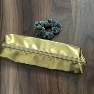  Pennskrin och Scrunchie  - Säljer ett guldfärgat pennskrin och en silverglittrig Scrunchie. Pennskrinet har guldfärgad dragkedja och båda är i bra skick. 