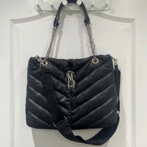 Svart quiltad axelväska från Steve Madden - Snygg svart quiltad axelväska från Steve Madden med kedjedetaljer och logotyp i silver. Väskan har både handtag och en justerbar axelrem för flexibilitet. Perfekt för att bära dina dagliga nödvändigheter med stil.