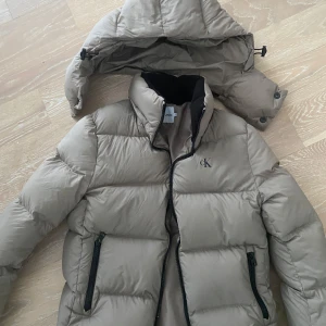 Beige dunjacka från Calvin Klein - Snygg beige dunjacka från Calvin Klein med avtagbar huva och dragkedja framtill. Jackan har två praktiska fickor med dragkedjor och en diskret CK-logga på bröstet. Använd fåtal gånger