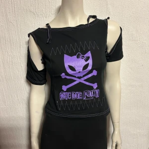 Svart topp med lila tryck S/M - Cool svart topp med ett lila katt- och korsbenstryck. Toppen har en unik design med avklippta axlar och knytband vid axlarna. Perfekt för en edgy stil.