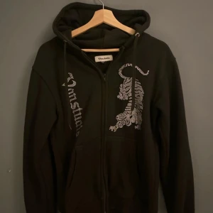 Svart hoodie med tigerdesign från Von Studio - Snygg svart hoodie från Von Studio med dragkedja och en cool tigerdesign i rhinestone på framsidan. Den har en justerbar huva med snören och långa ärmar. Perfekt för en avslappnad stil.
