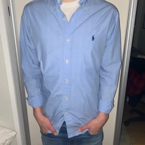 Blå skjorta från Ralph Lauren - Säljer en klassisk blå skjorta från Ralph Lauren med broderad logga på bröstet. Skjortan har långa ärmar och knappar framtill. Perfekt för en stilren look.