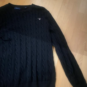 Svart kabelstickad tröja från Gant - Säljer en stilren svart kabelstickad tröja från Gant. Tröjan har en klassisk design med rund hals och långa ärmar. Perfekt för kyligare dagar.