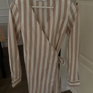 Randig omlottklänning  -  randig omlottklänning i  beige och vit. Klänningen har långa ärmar och ett knytband i midjan