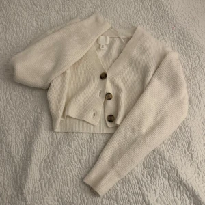 Vit kofta från H&M - Säljer en stilren vit kofta från H&M. Den har en croppad passform med stora knappar framtill och långa ärmar. Perfekt för lager-på-lager-stil eller som ett lättare plagg under kyligare dagar. I storlek S. 