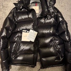 Svart dunjacka från Moncler - Snygg och varm svart dunjacka från Moncler med glansig finish. Jackan har en hög krage och dragkedja framtill samt praktiska fickor med dragkedjor. Perfekt för kyliga dagar.
