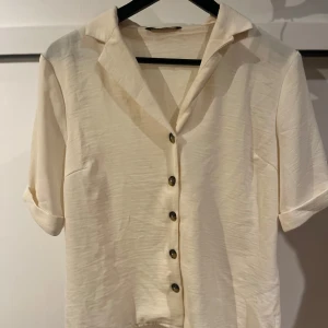 Krämfärgad blus med knappar från Only - Elegant krämfärgad blus med korta ärmar och knappar framtill. Perfekt för en stilren look. Stor i storleken. 