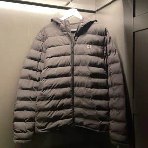 Svart pufferjacka från Fred Perry - Snygg svart pufferjacka från Fred Perry med dragkedja och huva. Perfekt för kyliga dagar med sin quiltade design och långa ärmar. Den har en stilren look med Fred Perry-loggan på bröstet.