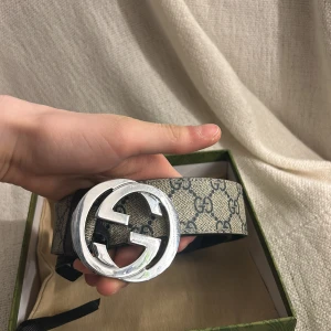 Mönstrat bälte med silverdetaljer - Snyggt bälte från Gucci med ett klassiskt mönster i beige och grått. Bältet har en stor, rund spänne i silver med märkets logotyp. Perfekt för att ge en lyxig touch till din outfit.