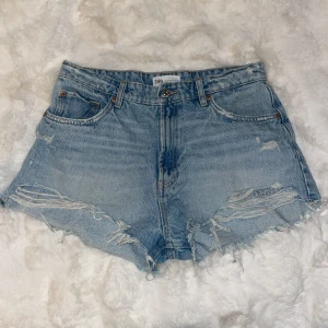 Ljusa jeansshorts från Zara - Säljer ett par ljusa jeansshorts från Zara med slitna detaljer och fransig kant. De har en klassisk femficksdesign och dragkedja. Perfekta för en avslappnad sommarlook! Säljer pga förstora.