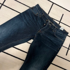 Mörkblå jeans från Carhartt - Snygga mörkblå jeans från Carhartt med klassisk femficksdesign och knappgylf. Perfekta för en avslappnad stil med en robust känsla. Märkeslogga på bakfickan ger en extra touch. 