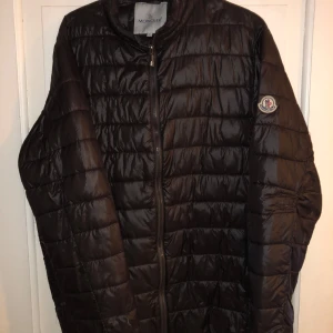 Svart dunjacka från Moncler - Säljer en stilren svart dunjacka från Moncler med dragkedja och deras ikoniska logotyp på ärmen. Jackan har en quiltad design och är perfekt för varmare väder. Passar perfekt till våren som är runt hörnet!