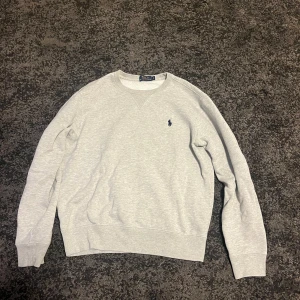 Grå tröja från Polo Ralph Lauren - Säljer en stilren grå tröja från Polo Ralph Lauren med deras ikoniska logga broderad på bröstet. Tröjan har långa ärmar och en klassisk rund halsringning. Perfekt för en avslappnad look.