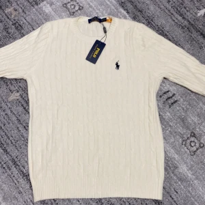 Vit stickad tröja från Ralph Lauren - Säljer en stilren vit stickad tröja från Ralph Lauren med det klassiska logotypbroderiet på bröstet. Tröjan har en rund halsringning och långa ärmar, perfekt för kyligare dagar. Ett måste i garderoben för den som älskar tidlös design.