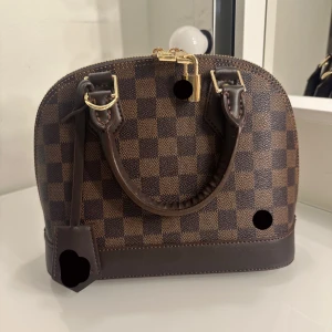 Brun rutig handväska - Snygg brun rutig handväska med guldfärgade detaljer och dragkedja. Väskan har ett klassiskt rutmönster och avtagbar axelrem. Perfekt för att ge en touch av elegans till din outfit.