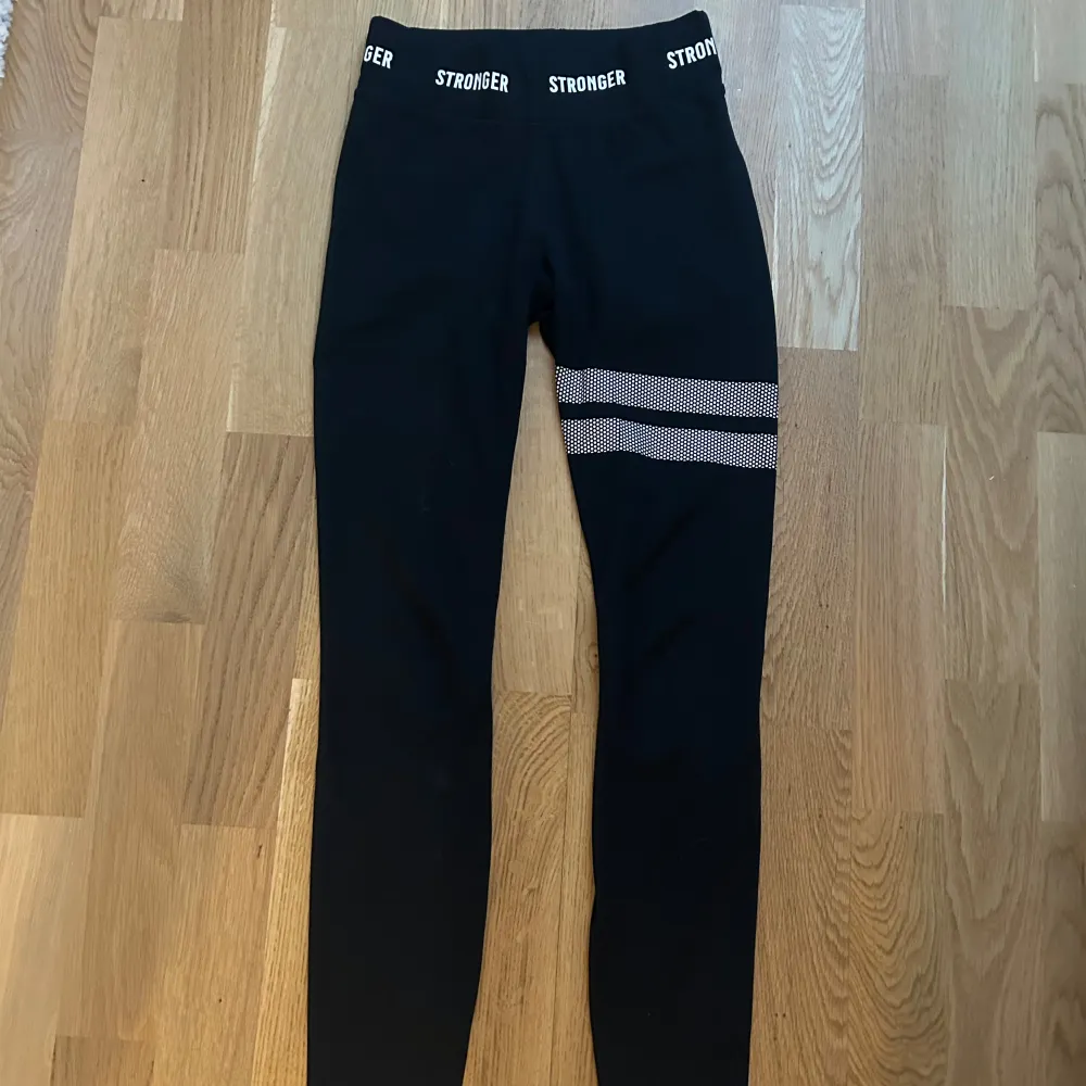 Snygga svarta leggings från Stronger med elastisk midja och ett coolt randigt mönster på ena benet. Perfekta för träning eller en avslappnad dag. De är stretchiga och bekväma, vilket gör dem till ett måste i garderoben.. Farkut & Housut.