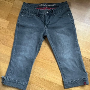 Grå capribyxor från Esprit - Snygga grå capribyxor från Esprit med knappar vid bensluten och en cool röd mönstrad insida. Perfekta för en avslappnad stil med en touch av detaljer. De har en klassisk femficksdesign och en bekväm passform.