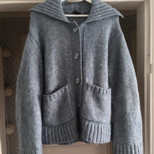Gråblå stickad kofta med knappar - Mysig gråblå stickad kofta med stora knappar och två praktiska fickor framtill. Den har en bred krage och ribbade detaljer vid ärmslut och nederkant. Perfekt för kyliga dagar. 