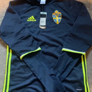 Mörkblå långärmad fotbollströja från Adidas - Snygg mörkblå långärmad fotbollströja från Adidas med Sveriges landslagsemblem på bröstet. Tröjan har neongula detaljer och Adidas-logga.