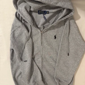 Grå hoodie från Polo Ralph Lauren - Säljer en stilren grå hoodie från Polo Ralph Lauren med dragkedja och justerbar huva. Den har en broderad logotyp på bröstet och är perfekt för en avslappnad look. Passar bra till både jeans och träningsbyxor. M/S storlek.