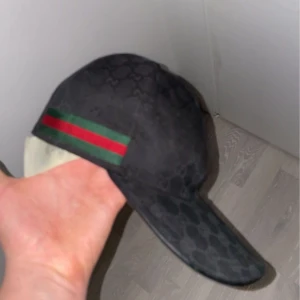 Svart keps med mönster och ränder - VILL BYTA MOT SAMMA I SVART ELR BLÅ MÅSTE VARA XS.   Snygg svart keps med diskret mönster och ikoniska gröna och röda ränder på sidan. Justerbar rem baktill för perfekt passform. Perfekt för att ge din outfit en stilfull touch. 