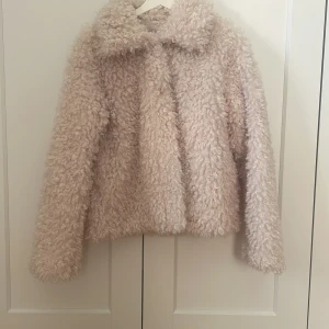 Beige teddyjacka - Mysig beige teddyjacka med långärmad design. Perfekt för att hålla sig varm och stilren under kyliga dagar. Jackan har en fluffig textur som ger en trendig look. Har inga defekter och har knappar för att knäppa ihop den. Är inte fluffig på insidan men du blir inte kall när du har på dig den. Har fickor på sidorna men dom syns i på bilden