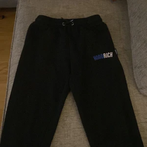 Svarta mjukisbyxor från Hoodrich - Säljer ett par svarta mjukisbyxor från Hoodrich med elastisk midja och snörning. Byxorna har en avslappnad passform och logotypen 'Hoodrich' tryckt i vitt och blått på sidan. Perfekta för en bekväm stil.