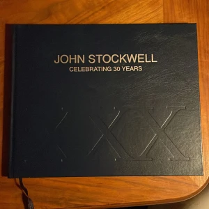 John Stockwell: Celebrating 30 Years - En bok som firar John Stockwells 30-åriga karriär med konstverk och texter. Boken har en mörkblå omslag med guldtext och innehåller färgrika landskapsmålningar och reflektioner från galleridirektören Zola Solamente.