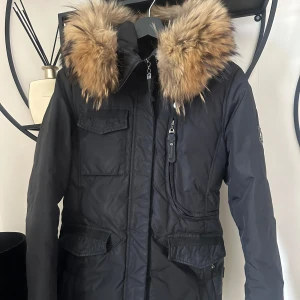 Svart Hollies vinterjacka med äkta päls - Snygg svart parkajacka med stor, fluffig pälsdetalj runt kragen. Jackan har flera praktiska fickor och dragkedja framtill. Jackan kommer från märket Hollies och är i storlek 38