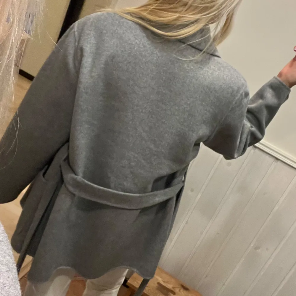 Säljer en stilren grå kappa från Stockh LM. Den har en klassisk design med knytskärp i midjan och stora fickor framtill. Perfekt för kyligare dagar med sin långa ärm och mjuka ullblandning. En tidlös favorit i garderoben!. Takit.