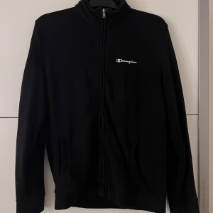 Svart kofta från Champion - Säljer en svart kofta från Champion med dragkedja framtill. Jackan har en enkel design med Champion-loggan på bröstet. 