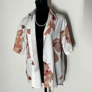 Vit blommig kortärmad skjorta - Snygg vit skjorta med stora blommönster i rosa och beige toner. Skjortan har korta ärmar och knappar framtill, perfekt för en avslappnad stil.