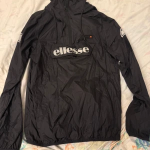 Svart vindjacka från Ellesse - Säljer en svart vindjacka från Ellesse med stor logga på framsidan. Jackan har en huva med dragsko och långa ärmar. Perfekt för blåsiga dagar.