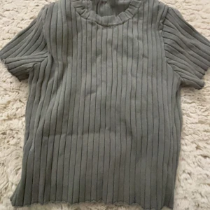 Grå ribbad tröja - Säljer en stilren grå ribbad tröja med korta ärmar. Perfekt för en avslappnad look. Tröjan har en rund halsringning och är gjord i ett mjukt material.