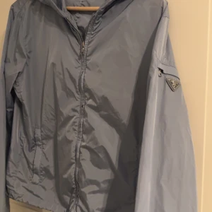 Prada windbreaker  - Säljer en stilren blå vindjacka från Prada med dragkedja och huva. Jackan har en ficka med dragkedja på ärmen och en diskret logotyp. Perfekt för blåsiga dagar. Jackan har ett märke på höger arm som ev går bort i kemtvätt, därav det låga priset! Storlek 44=XS