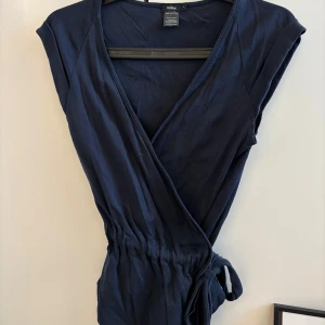 Mörkblå omlottopp från Filippa K - Säljer en stilren mörkblå omlottopp från Filippa K i 100% bomull. Toppen har en knytning i midjan som du kan reglera själv den har och korta ärmar.