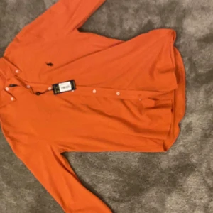 Orange långärmad skjorta från Ralph Lauren - Snygg orange långärmad skjorta från Ralph Lauren med klassisk krage och knappar framtill. Skjortan har en broderad logga på bröstet och är perfekt för en stilren look. Passar bra till både jeans och chinos. Helt ny pris lappen sitter kvar